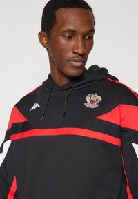 Črna jopa s kapuco z rdečimi in belimi črtami, z izrazitim vezenim logotipom. Material je videti mehak, z udobnim krojem in nastavljivim vrvico za zategovanje.