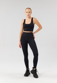 Melns sporta kultūrkrekls un augsta vidukļa sporta bikses no elastīga materiāla, kopā ar melnām sporta kurpēm ar reljefām zolēm.