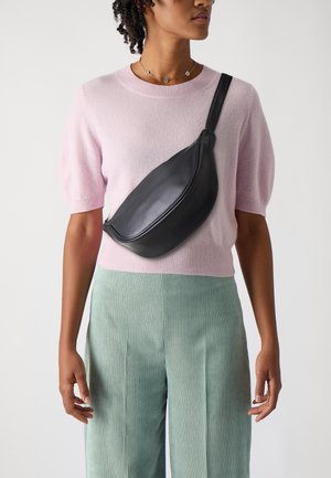 Černá kožená crossbody taška s zakřiveným designem, nošená přes světle růžový pletený svetr a zelené žebrované kalhoty. Hladká textura, minimalistický styl.