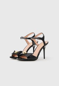 Black patent leather high-heeled sandals with open toes, adjustable ankle strap, and gold heart accent on the bow detail.
