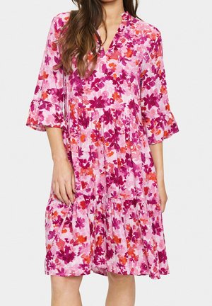Femme portant une robe rose ample longueur genou avec des motifs floraux violets et orange et des manches trois-quarts à volants.