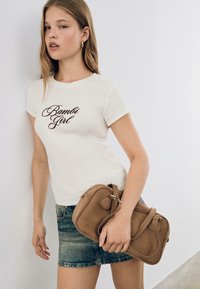 Weißes Baumwoll-T-Shirt mit "Bambi Girl" in brauner Schreibschrift, kombiniert mit einem blauen Denim-Rock und einer hellbraunen Wildleder-Handtasche. Einfaches und lässiges Design.