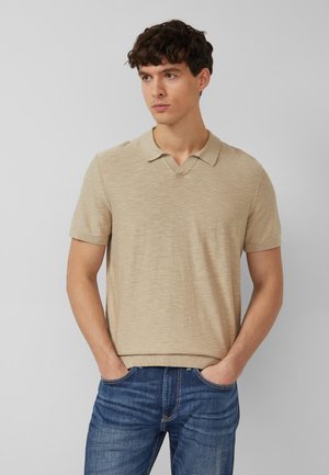 Giovane uomo con capelli scuri e ricci che indossa una polo beige a maniche corte in maglia e jeans blu, con le mani in tasca, che guarda a sinistra.