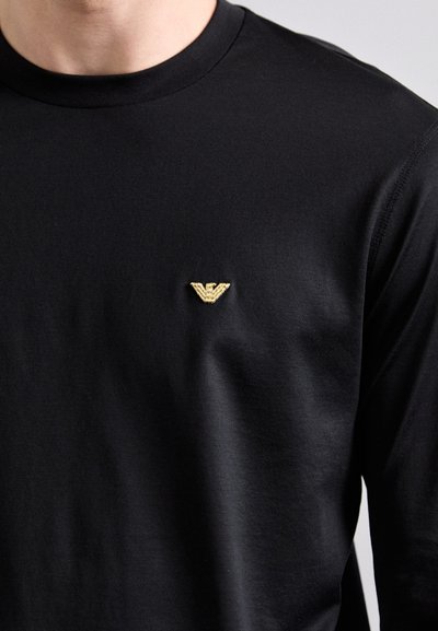 T-shirt noir à manches longues en tissu lisse, avec un petit logo doré sur le côté gauche de la poitrine. Design simple et ajusté.
