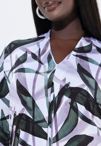 Detailní záběr osoby nosící top s výstřihem do V s motivy listů v zelené, černé a levandulové barvě, stříbrné kruhové náušnice a lesklé rty.