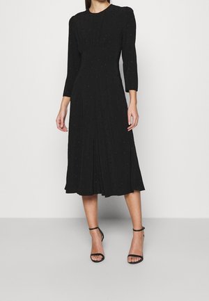 Musta midimekko, jossa on kolme neljännestä hihaa, teksturoitu kangas, vartalonmyötäinen yläosa ja liehuva helma. Yhdistetty mustiin remmikkäisiin korkokenkiin.