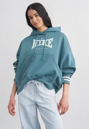 Jeune femme aux longs cheveux foncés portant un sweat à capuche Nike bleu sarcelle et un jean bleu clair, debout devant un fond clair uni.