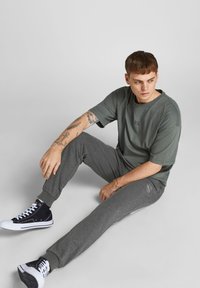 Graue Baumwoll-Jogginghosen mit einem gerippten Bündchen, kombiniert mit einem lockeren olivgrünen T-Shirt mit einem kleinen Grafikdruck. Schwarze High-Top-Sneaker.