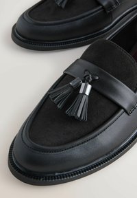Sorte loafers lavet af glat læder og ruskind, med en dekorativ kvast og syet detaljer langs sålen.
