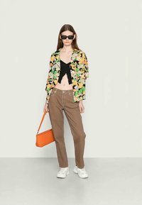 Camisa de manga curta com estampa floral, top cropped preto, calças castanhas de perna reta, ténis brancos, mala laranja e óculos de sol escuros.