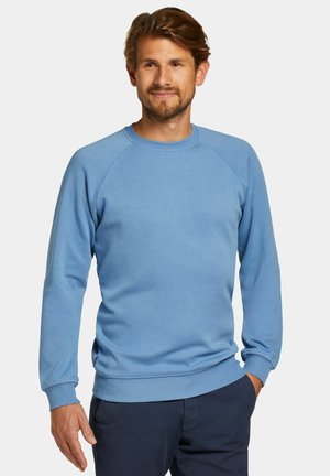 Uomo con capelli castano chiaro indossa una felpa azzurro chiaro a maniche lunghe e pantaloni blu scuro, in piedi con una mano in tasca davanti a uno sfondo semplice.