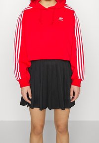 Röd kort hoodie med vita ränder på ärmarna och liten logotyp, i kombination med en svart plisserad kjol. Slät tygtextur, sportig design.