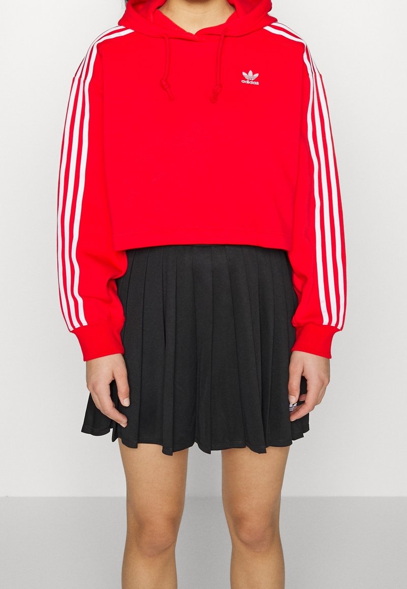 Röd kort hoodie med vita ränder på ärmarna och liten logotyp, i kombination med en svart plisserad kjol. Slät tygtextur, sportig design.