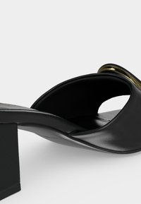 Μαύρο δερμάτινο mule με ανοιχτά δάχτυλα, τακούνι block και χρυσή μεταλλική λεπτομέρεια στην κορυφή, εμφανιζόμενο από πλευρική γωνία σε λευκό φόντο.