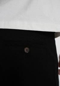 Nahaufnahme einer schwarzen Hosentasche mit Knopf und dezent gesticktem Logo unter dem Saum eines weißen Hemdes.