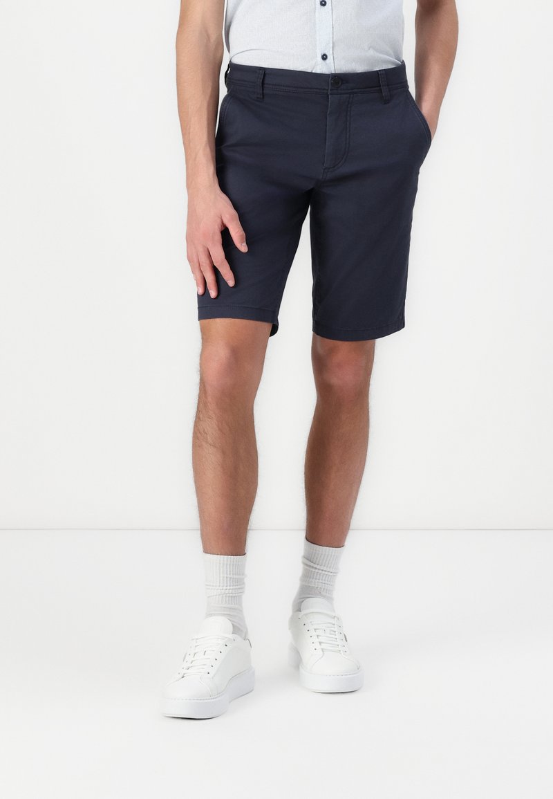 Shorts bleu marine en mélange de coton, jusqu'aux genoux avec poches latérales. Une texture lisse et une coupe ajustée, assortis à des baskets blanches et des chaussettes grises.
