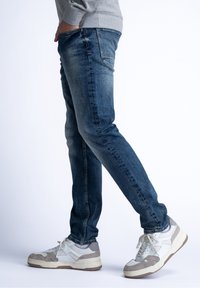 Tmavě modré džínové kalhoty s úzkým střihem, s jemným vyblednutím, viditelnými švy a detailem zadní kapsy. Doplněné bílými teniskami.