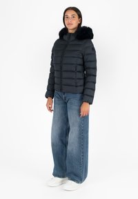 Giubbotto piumino nero con un design trapuntato testurizzato e un bordo in pelliccia sintetica. Indossato sopra jeans blu larghi e sneakers bianche.