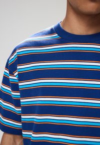 Blå stribet t-shirt med horisontale striber i lyseblå, mørkblå, hvid og orange. Standard crewneck-design med blød tekstur.