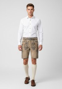 Lederhosen-shorts i lysebrun med detaljeret mørk broderi, parret med en hvid skjorte med knapper og cremefarvede knæstrømper. Brune sko fuldender looket.