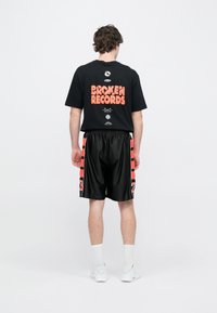 Μαύρο μπλουζάκι με πορτοκαλί γραφικά που γράφουν «BROKEN RECORDS». Μαύρο σορτς με κόκκινες λεπτομέρειες και τον αριθμό «23» στο πλάι. Λευκά αθλητικά παπούτσια.