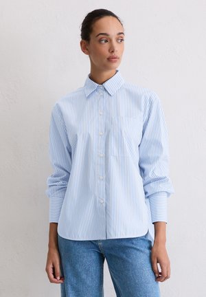 Camicia azzurra chiaro con righe bianche verticali, dotata di colletto, maniche lunghe e taschino sul petto. Indossata con jeans a vita alta.