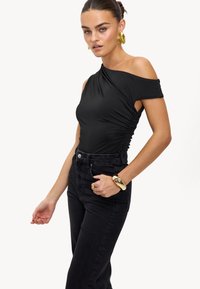 Bodysuit noir à épaule unique avec un design froncé et une découpe sur le côté, associé à un jean noir taille haute. Boucles d'oreilles et bracelet en or remarquables.