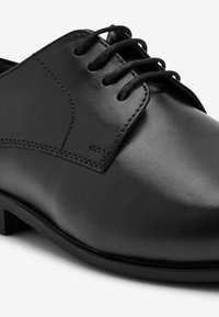 Chaussure habillée en cuir noir avec une finition lisse, bout rond, six œillets et lacets noirs. Présente un subtil détail de couture le long du panneau.
