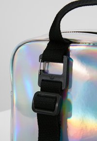 Bolsa de PVC holográfica con una correa de nylon negra y un cierre de plástico. Forma redondeada con superficie reflectante que exhibe colores iridiscentes.