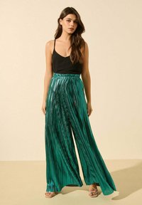 Pantalons larges plissés verts avec une texture brillante, taille élastique et design fluide, associés à un caraco noir et des sandales nues.