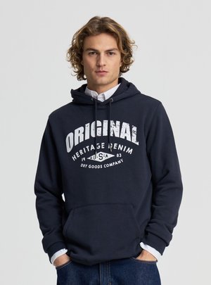 Giovane uomo con capelli ricci che indossa una felpa con cappuccio blu navy con la scritta bianca "ORIGINAL Heritage Denim USA" e jeans, mani nelle tasche, sfondo neutro.