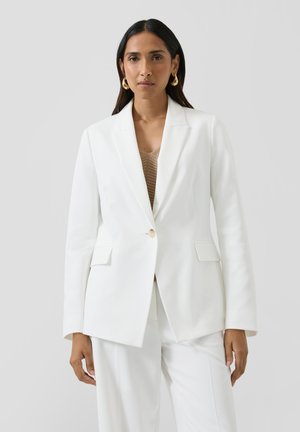 Femme portant un blazer blanc cintré et un pantalon assorti, debout devant un fond gris clair uni.
