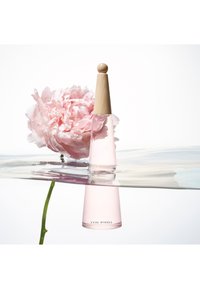 Transparent glass parfymeflaske med trelokk, fylt med lys rosa væske, delvis nedsenket i vann ved siden av en rosa pion.