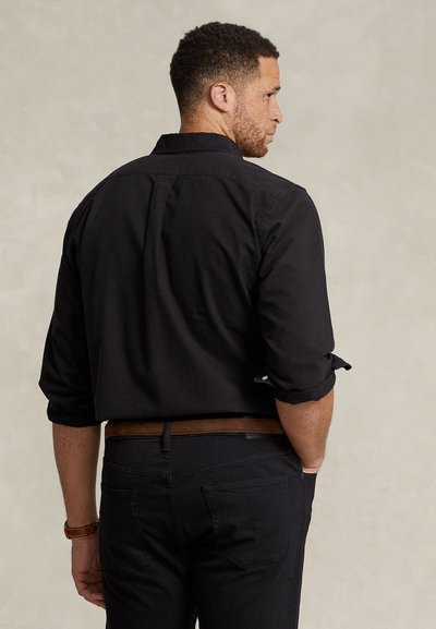 Man wearing a black long-sleeve shirt with sleeves rolled up, black pants, brown belt, and brown watch, facing away against a neutral background.