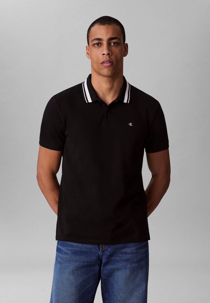 Black polo shirt with white striped collar, short sleeves, and a small logo on the chest, paired with blue jeans. Smooth fabric texture.