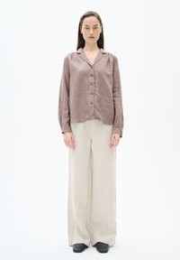 Knoopshirt van mauve linnen, met lange mouwen en een zachte textuur, gecombineerd met ruime beige linnen broek en zwarte schoenen.