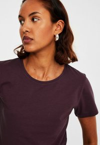 Pflaumefarbener Rundhalsausschnitt T-Shirt aus weichem Stoff, mit kurzen Ärmeln und glatter Textur. Das Model trägt silberne ovale Ohrringe.