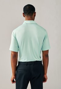 Ljusgrön pikétröja med korta ärmar, i texturerat tyg och med klassisk krage, kombinerad med mörkblå byxor. Bakifrån.