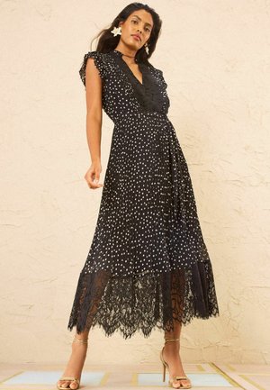 Frau trägt ein schwarz-weiß gepunktetes Kleid mit Spitzenbesatz, steht vor einer strukturierten beigen Wand, geschmückt mit Blumenohrringen.