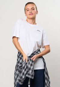 Ung kvinde justerer sort-hvid ternet skjorte bundet om livet, iført hvid t-shirt og mørke bukser mod en ensfarvet baggrund.