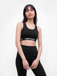 Joven mujer con un sujetador deportivo negro con el logo "Urban Classics" y mallas de cintura alta a juego, posando frente a un fondo blanco liso.