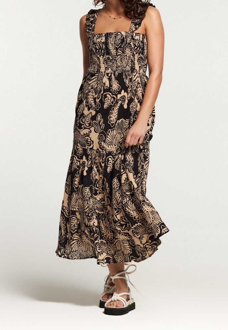 Maxi dress in black with beige floral print, featuring ruffled straps, a smocked bodice, and a flared skirt. Paired with white platform sandals.