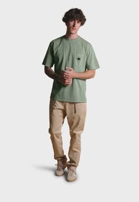 Světle zelené bavlněné tričko s kapsou na hrudi a černým logem slona, v kombinaci se světle béžovými kalhotami a hnědými teniskami.