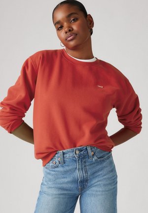 Junge Frau mit dunkler Haut und Haaren zu einem Dutt gebunden, die goldene Creolen, ein rotes Levi's Sweatshirt und hochgeschnittene blaue Jeans trägt, vor einem schlichten Hintergrund.