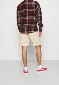 Chemise à carreaux rouge et noir, shorts beiges et baskets rouges avec des accents blancs. La chemise a des manches longues et une coupe décontractée.