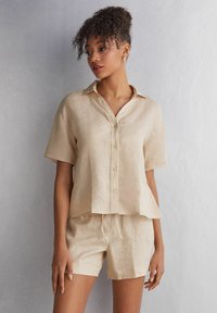 Beige lněná košile s krátkým rukávem, zapínáním na knoflíky a volným střihem, doplněná o šortky na šňůrku, které barevně ladí. Hladká textilie, ležérní design.
