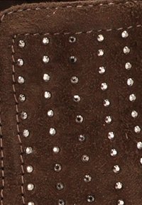 Tejido de ante marrón con pequeños clavos de plata distribuidos de manera uniforme en filas verticales y costura visible a lo largo del borde.
