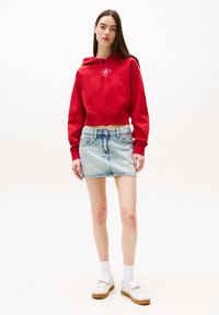 Hoodie rosso cropped con logo frontale, abbinato a una mini gonna di jeans azzurri e sneakers bianche con suole marroni. Texture lisce in tutto.