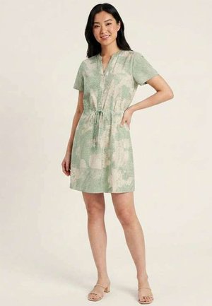 Femme debout, main sur la hanche, vêtue d'une robe à manches courtes, longueur genou, à motifs verts et beiges, et de sandales beiges aux orteils ouverts.