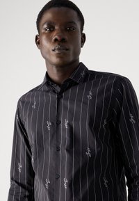 Camicia nera con bottoni e strisce verticali bianche, impreziosita da ricami. Caratterizzata da un colletto strutturato e una forma aderente. Texture liscia.